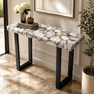 May include: A rectangular console table featuring a top made of polished, multi-tonal white and grey agate stone slices with dark veining. The table is supported by two sturdy, black metal U-shaped legs that provide a stable base for the heavy stone top.