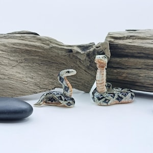 May include: Two ceramic snake figurines with detailed patterns in black, white, and tan. One is coiled, the other upright. The background includes weathered wood and smooth, dark grey stones, creating a naturalistic display.