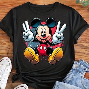 May include: Black t-shirt featuring a colorful graphic of Mickey Mouse making peace signs. Mickey is in his classic red shorts, yellow shoes, and white gloves. The background has red and blue accents with small stars.