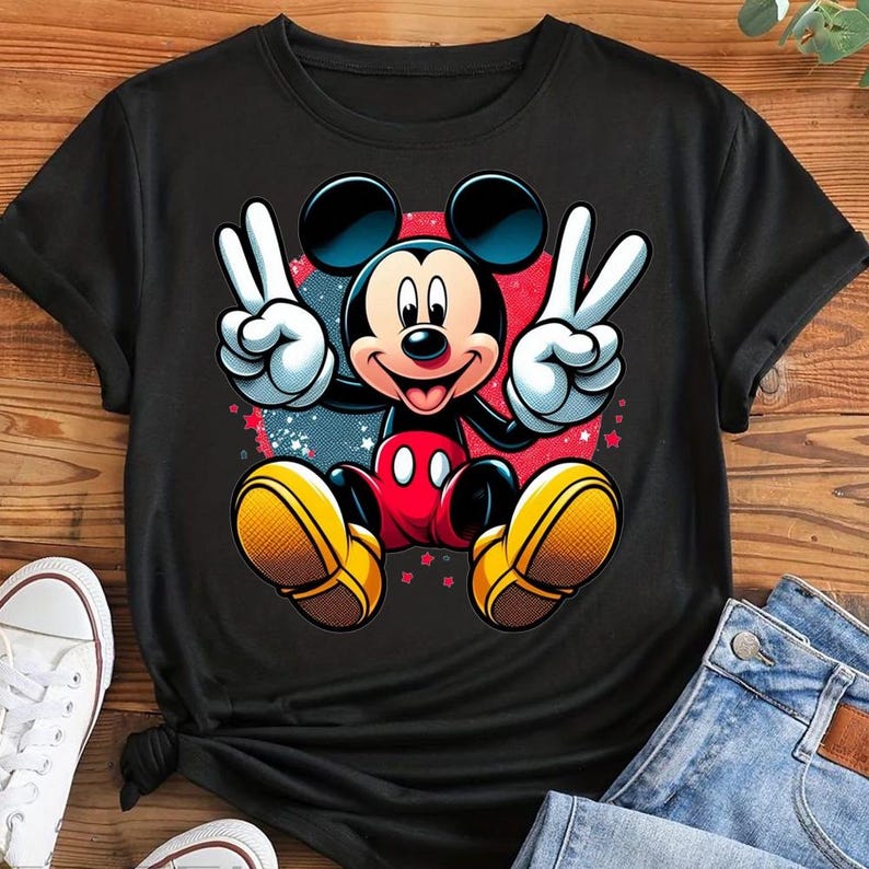May include: Black t-shirt featuring a colorful graphic of Mickey Mouse making peace signs. Mickey is in his classic red shorts, yellow shoes, and white gloves. The background has red and blue accents with small stars.