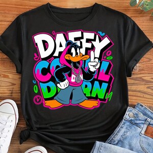 May include: Black t-shirt featuring a cartoon Daffy Duck character in a pink hoodie, silver chain, and blue pants. The shirt has the words "DAFFY COOL DARN" in a graffiti-style design with bright colors.