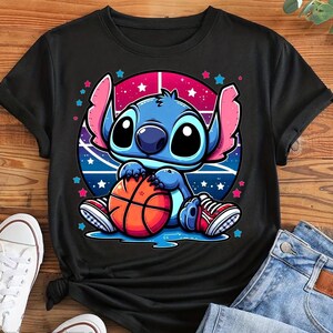 May include: Black t-shirt featuring a cartoon character holding a basketball. The character is blue with large eyes and pink ears, set against a pink and blue background with stars. The basketball is orange and brown. The character is wearing red and white sneakers.