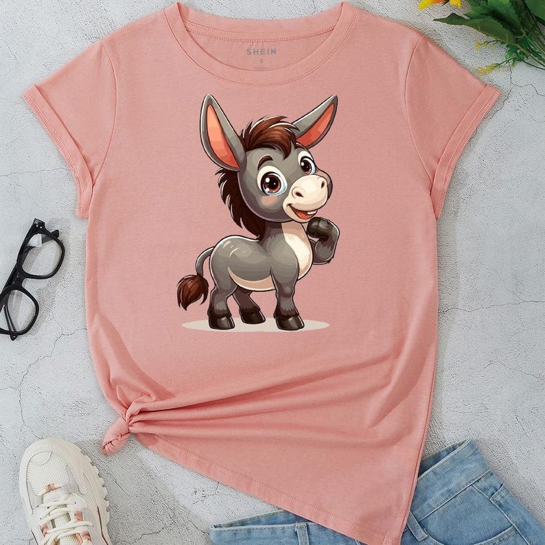 May include: A light pink t-shirt featuring a cartoon donkey illustration. The donkey is gray with brown hair and a friendly expression. The shirt has rolled-up sleeves and the brand name "SHEIN" is printed on the top.