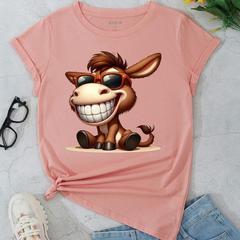 May include: A light pink t-shirt featuring a cartoon donkey wearing sunglasses and a big smile. The donkey is brown with a white smile and is sitting down. The shirt has rolled-up sleeves and a knot at the bottom.