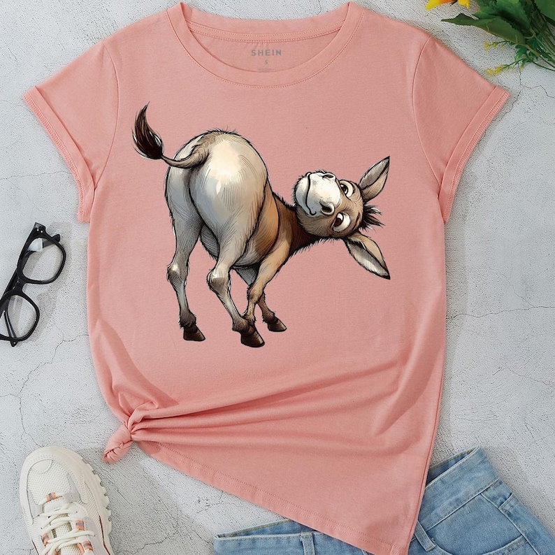 May include: A light pink t-shirt featuring a cartoon donkey illustration. The donkey is depicted in shades of brown, tan, and white, with its head tilted and looking upwards. The shirt has rolled sleeves and a knot detail at the bottom.