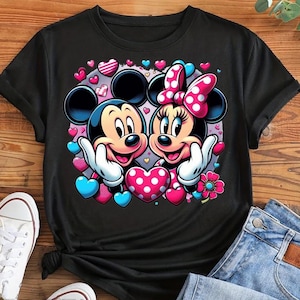 May include: Black t-shirt featuring Mickey and Minnie Mouse embracing, surrounded by pink, blue, and white hearts. Minnie wears a pink polka-dot bow. The design includes a pink flower and white gloves.