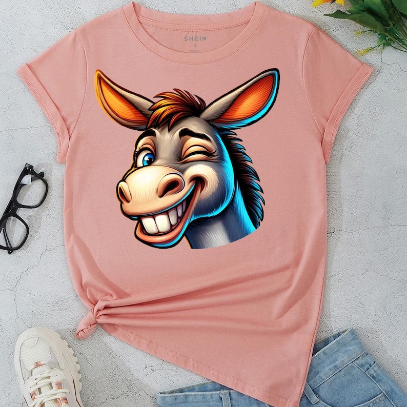 May include: A light pink t-shirt featuring a cartoon donkey winking and smiling. The donkey has gray fur, blue eyes, and large orange ears. The shirt has rolled-up sleeves and a knot at the bottom.