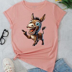 May include: A light pink t-shirt featuring a cartoon donkey with a large grin and pointing finger. The donkey has brown fur, a blue and brown eye, and large teeth. The shirt has rolled sleeves and a knotted hem.