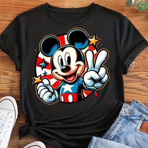 May include: Black t-shirt featuring a colorful graphic of Mickey Mouse. Mickey is depicted with a patriotic theme, wearing a red, white, and blue outfit and giving a peace sign. The design includes stars and a red, white, and blue circular background.