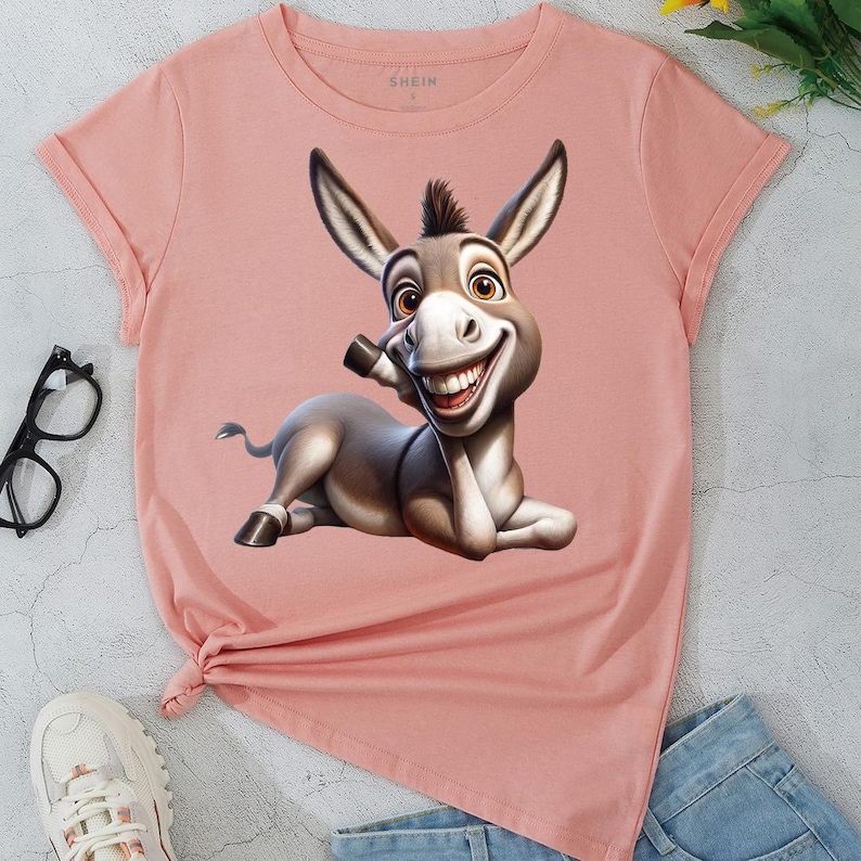 May include: A light pink t-shirt featuring a cartoon donkey with a large smile. The donkey is lying down and has brown and gray fur. The shirt has rolled-up sleeves and a knot at the bottom.