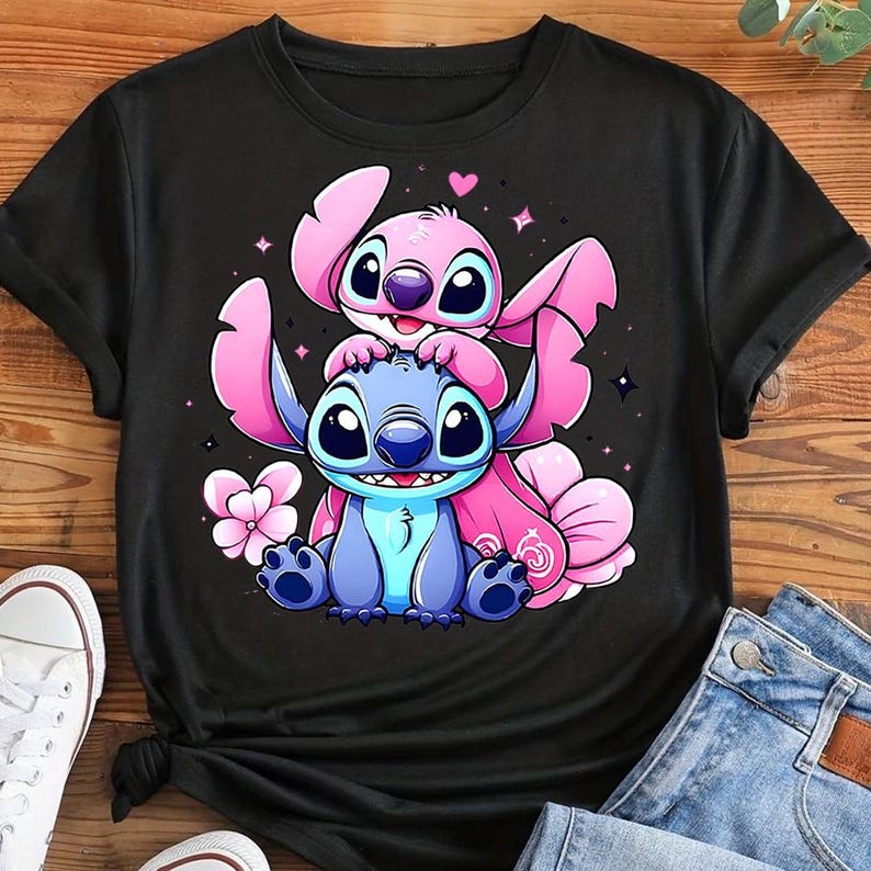 May include: Black t-shirt featuring a cartoon illustration of two characters, one blue and one pink, with large eyes and ears. The pink character is on top of the blue one. Pink flowers and stars surround the characters.