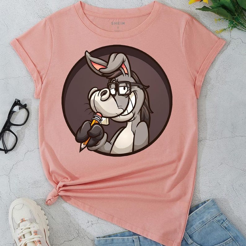May include: A light pink t-shirt featuring a cartoon donkey wearing glasses and holding a pencil. The donkey is inside a dark brown circle. The shirt has rolled-up sleeves and a knot at the bottom.