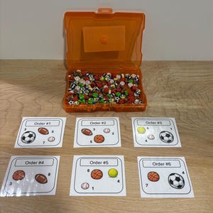 May include: An orange plastic container filled with miniature sports balls, including soccer, basketball, baseball, and football. Six cards with the text "Order #" and sports ball illustrations are arranged below the container.