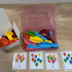 May include: A set of colorful balloons in a pink plastic container and a white box. The balloons are in various colors, including red, yellow, orange, pink, green, and blue. Several cards with balloon illustrations are also present.