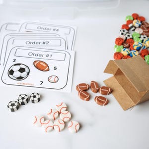 May include: A set of small sports-themed erasers including soccer balls, footballs, and baseballs, with a brown paper box and several printed order cards labeled Order #1, Order #2, and Order #4, each displaying specific quantities of each ball type.