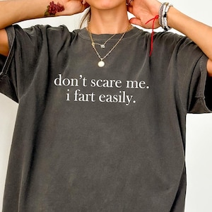 May include: Dark gray t-shirt with the text "don't scare me. i fart easily." in white. The shirt is a relaxed fit and has short sleeves. The word "PEPPER" is printed in the bottom right corner.