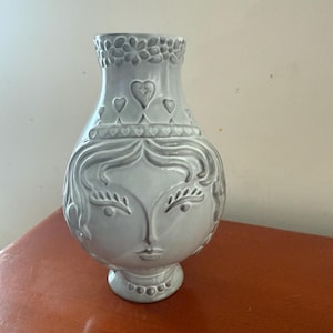 Può includere: Un vaso in ceramica grigia con un disegno di volto. Il vaso presenta il volto di una donna con occhi, naso e labbra dettagliati. La parte superiore del vaso ha un motivo floreale e il corpo ha accenti di cuore e fiori. Il vaso è su una superficie marrone.