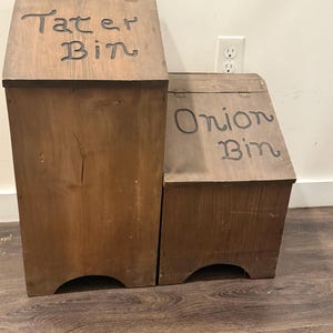 May include: Two wooden storage bins with hinged lids. The taller bin is labeled "Tater Bin" and the smaller one is labeled "Onion Bin" in a handwritten style. Both bins are made of stained wood and have curved cutouts at the base.