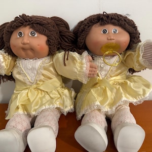 May include: Two vintage Cabbage Patch Kids dolls. Each doll has brown yarn hair, rosy cheeks, and blue eyes. They wear yellow satin dresses with lace trim and white lace-trimmed stockings. One doll has a pacifier.