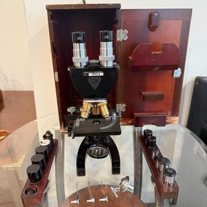 Puede incluir: Un microscopio negro antiguo con componentes de latón y dos oculares, expuesto abierto en una caja de madera. Lentes y herramientas adicionales están dispuestas sobre una superficie de vidrio, lo que sugiere un tema científico o educativo.