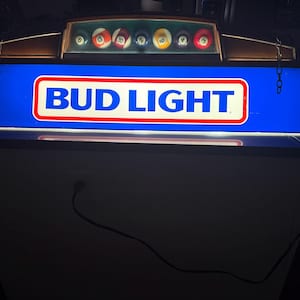 May include: A blue Bud Light sign with a white and red border. Above the sign is a display of billiard balls. The sign is mounted on a white cabinet with a power cord.