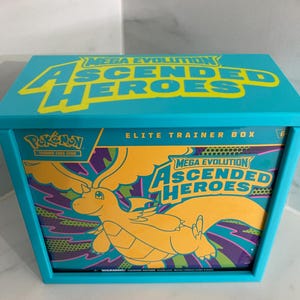 Puede incluir: Una caja de entrenador de élite turquesa para el juego de cartas coleccionables Pokémon. La caja presenta el texto "Mega Evolution Ascended Heroes" en amarillo y un gráfico de una criatura amarilla con forma de dragón. La caja es rectangular con tapa.