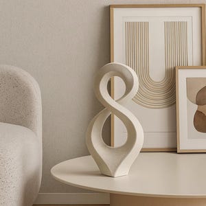Op de afbeelding: Een crèmekleurige abstracte sculptuur in de vorm van een oneindigheidssymbool staat op een ronde, gebroken witte tafel. Ingelijste kunst en een stoel zijn op de achtergrond. De kamer heeft een neutraal kleurenpalet.