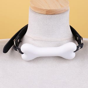 May include: A black leather collar with a white bone-shaped centerpiece. The collar is displayed on a mannequin with a wooden top. The bone is a solid white color and the leather is black.