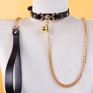 May include: Black leather collar with gold-colored hardware, a small bell, and a matching gold chain leash. The collar is studded and has a buckle closure. The leash has a loop handle.