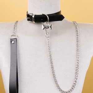 May include: Black leather choker with a silver buckle and a bone-shaped charm. A silver chain and a matching black leather handle are attached. The choker is displayed on a mannequin.