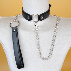 May include: Black leather choker with a heart-shaped center ring and a silver chain leash. The choker has a matching black leather handle. The design is displayed on a white mannequin.