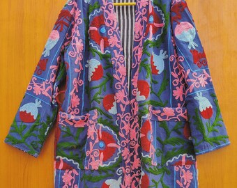 May include: A long, open-front coat in a vibrant blue hue, adorned with intricate floral embroidery in red, pink, green, and white. The coat features two pockets and a striped interior lining, showcasing a bohemian aesthetic.