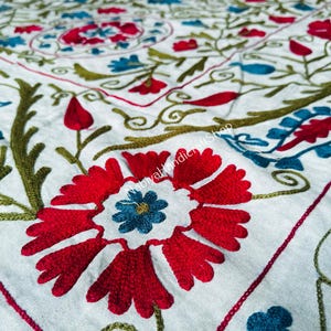 Peut inclure: Un textile en tissu blanc orné de motifs floraux brodés à la main avec des fils rouge vif, bleu profond et vert olive. Le design présente de grandes fleurs stylisées, des vignes entrelacées et des bordures géométriques avec le texte NobiyaHandicraftShop.