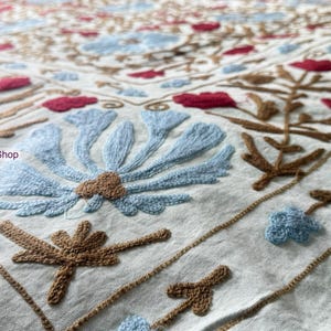 Peut inclure: Gros plan sur un textile blanc avec une broderie détaillée. Le motif présente de grandes fleurs bleu clair, des accents floraux rouges et des tiges et feuilles marron. La broderie est complexe et semble faite à la main.