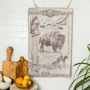 May include: A brown and white kitchen towel with a vintage illustration of a buffalo, Native American figures, and a horse and rider. The towel is hanging on a white brick wall with a wooden cutting board and a bowl of fruit in the foreground.