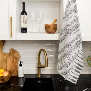 May include: A kitchen sink with a gold faucet, a white tiled backsplash, and a black countertop. There is a wooden cutting board, a bottle of wine, two wine glasses, a wooden bowl, and a dish of lemons on the counter. A white tea towel with a black musical note design hangs on the cabinet door.