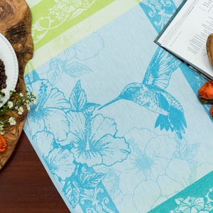 May include: A light blue and white patterned table runner featuring a hummingbird and hibiscus flowers. The runner is next to a wooden serving board with a white cup, coffee beans, and orange roses. A cookbook and wooden spoon are also present.