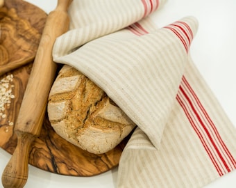 May include: A loaf of sourdough bread on a wooden cutting board, partially covered by a beige and red striped linen tea towel. The bread is on a wooden cutting board with a rolling pin and oats.
