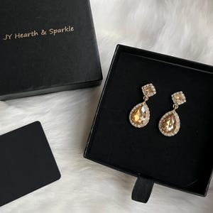 May include: A pair of gold-toned drop earrings with a square top and a teardrop-shaped bottom. The earrings are set with clear crystals and a large amber-colored stone. The earrings are displayed in a black box with the text "JY Hearth & Sparkle".