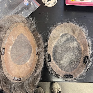 May include: Two grey hairpieces with a skin-toned base and attached clips. The hairpieces are on a dark surface, with a small decorative coaster with the number "1" visible. The hairpieces appear to be for hair replacement or styling.