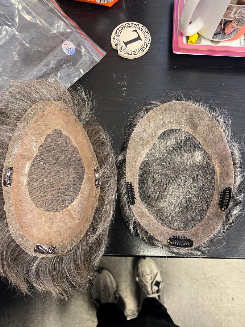 May include: Two grey hairpieces with a skin-toned base and attached clips. The hairpieces are on a dark surface, with a small decorative coaster with the number "1" visible. The hairpieces appear to be for hair replacement or styling.