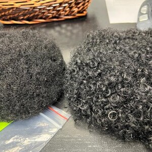 May include: Two dark grey afro hair pieces with different textures. One is tightly coiled, the other is more loosely curled. A wicker basket and a keyboard are in the background.