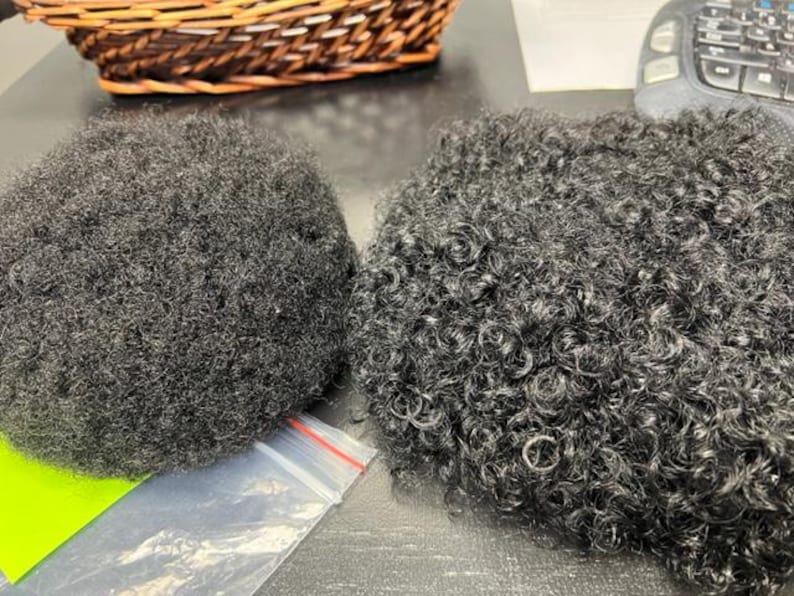 May include: Two dark grey afro hair pieces with different textures. One is tightly coiled, the other is more loosely curled. A wicker basket and a keyboard are in the background.