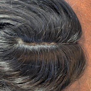 May include: Close-up of a dark hairpiece with a visible parting. The hair is dark with strands of grey, and the texture appears natural. The hairpiece is likely designed for cosmetic purposes.