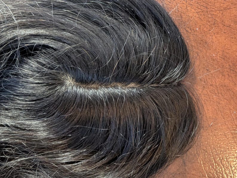 May include: Close-up of a dark hairpiece with a visible parting. The hair is dark with strands of grey, and the texture appears natural. The hairpiece is likely designed for cosmetic purposes.