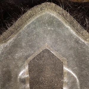 May include: Close-up of a hairpiece base, featuring a transparent, fine mesh material. The base is designed with a contoured shape, and the edges are reinforced with a darker, more durable material. Dark hair is visible beneath the base.