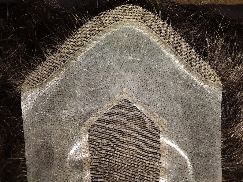 May include: Close-up of a hairpiece base, featuring a transparent, fine mesh material. The base is designed with a contoured shape, and the edges are reinforced with a darker, more durable material. Dark hair is visible beneath the base.