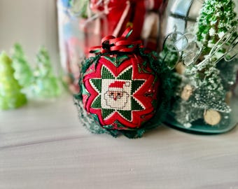 May include: A quilted fabric Christmas ornament featuring a central cross-stitch square of a Santa Claus face. The ornament is decorated with a red and green star pattern, dark green lace trim, and a red ribbon bow at the top, positioned near a glass jar.