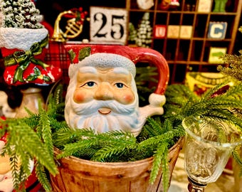 May include: A ceramic Santa Claus pitcher featuring a detailed face with a white beard, rosy cheeks, and a red hat brim. The handle is red, and the top rim is decorated with green holly leaves and red berries, nestled within a wooden basket.