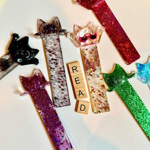 May include: A collection of cat-shaped resin bookmarks in various colors, including red, purple, green, and black, with glitter accents. The word "READ" is spelled out with wooden letter tiles.